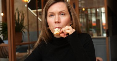 Güzel, şık bir kadın, restoranda oturmuş, bir parça tohumlu çiftlik ekmeği yiyor. Uzun kollu siyah gömlekli orta boy kadın. Susamlı taze çörek yiyor..