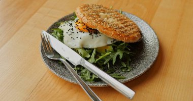 Kentsel kahvaltı susamlı simit kızarmış yumurta ve çatal ve bıçakla yeşillikler. Dengeli şehir brunch 'ı ya da öğle yemeği stili gri seramik tabakta servis ediliyor ve ahşap masada çelik çatal bıçak takımı var. Yüksek kalite fotoğraf