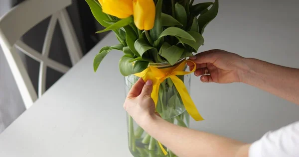 Female hands tie band bow on glass vase with fresh bunch yellow tulip. Beautiful bouquet with bright green leaves stands on table. High quality photo