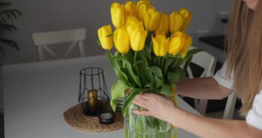 Uzun kahverengi saçlı kadın yemek masasına vazo ve sarı lale koydu. Beyaz tişörtlü kadın, ev masasını bir demet Hollanda çiçeğiyle dekore ediyor. Yüksek kalite 4k görüntü