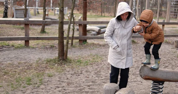 Anne oğluyla ahşap oyun parkında oynuyor ve yaylı kirişten kütüğe zıplamaya yardım ediyor. Beş yaşındaki çocuk, soğuk bir sonbahar gününde annesiyle dışarıda vakit geçirmekten hoşlanıyor..