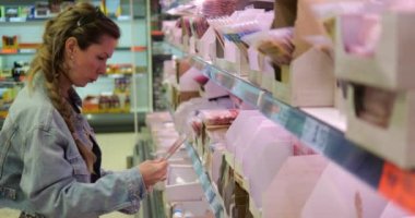 Şaşırmış kadın müşteri farklı marka soğuk et vakumlarını alıp kontrol ediyor. Kadın müşteri süpermarkette üst üste yenmiş et arıyor. Yüksek kalite 4k görüntü