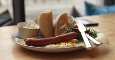 Kızarmış yumurta, yeşil salata, sosis, beyaz ekmek ve sosis. Lokantada pencerenin karşısındaki ahşap masada duran klasik kahvaltılı tabak. 4k görüntü.