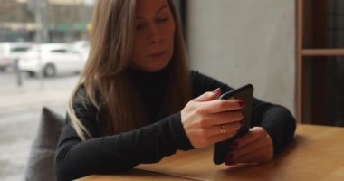 Ciddi bir şekilde konsantre olmuş düşünceli kadın şehir kafesindeki akıllı telefon ekranından okuyor. Genç kadın, telefon aygıtı ekranına yakından ve dikkatle bakıyor. Büyük pencerenin yanındaki masada hareketsiz oturuyor. 4k