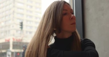 Genç ve güzel bir kadın, uzun kahverengi saçları şehir caddesine bakan pencerede otururken düzeltir. Siyahi balıkçı yakalı, kendine güvenen, şık bir kadının yakın plan fotoğrafı. Kamu binasında bekleyen saç modelini kontrol et. 4k