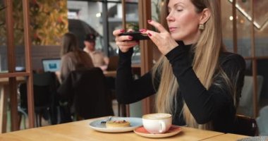 Güzel bir kadın kahve dükkanında tatlı ve kapuçino fotoğrafı çekiyor. Genç şık bakımlı kadın modern şehir kafesinde masada otururken telefonla kahve fotografı çekiyor. 4k