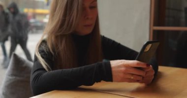 Genç bir kadın akıllı telefon ekranında yazı yazıyor ve restoranda oturmuş gülümsüyor. Kadın müşteri sosyal medyada zaman geçirir ve geçirir, haberlere bakar ve tepki verir ve yorumlara ve mesajlara cevap verir. 4k