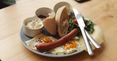 Kahvaltı tabağı yumurta, domatesli yeşil salata, sosis, çörek ve sos. Ahşap masada soslu ve çatal bıçaklı bir tabak iştah açıcı brunch yemeği. 4k görüntü.