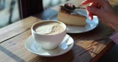 Kadın kahvaltıda kapuçino ile süt köpüğü yaprağını karıştırıyor. Ahşap tezgahta elinde kahve fincanı ve peynirli kek olan kadın. Cappuccino içeceği