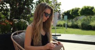 Büyük güneş gözlüklü kadın yaz parkında el çantasından pudra alıyor. Bayan, sokak kafesi terasında dinlenirken çantasında küçük bir ayna buluyor.