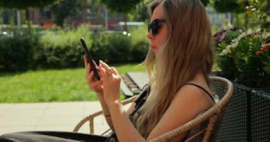Uzun sarı saçlı, omuz üstü ceketli kadın terasta otururken telefonda mesaj yazıyor. Dijital iletişimin görselleştirilmesi