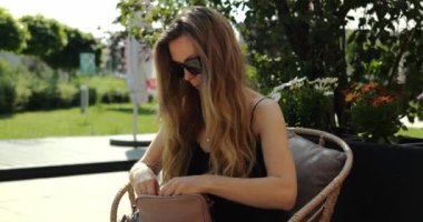Saçları dökülmüş şık bir kadın yaz parkında özel tasarım bir çantada makyaj malzemesi arıyor. Güneş gözlüklü bir kadın kafeterya terasında hasır koltukta oturuyor.
