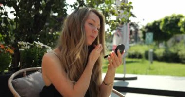 Uzun saçlı odaklanmış bir kadın, açık hava kafesindeki küçük aynaya bakarak göz makyajını ayarlıyor. Kadın gölgeli parkta ideal görünme konusunda endişeli