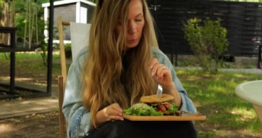 Sarışın, gevşek saçlı genç kadın gölgeli sokak terasında öğle yemeğinin tadını çıkarıyor. Bayan parkta rahatça otururken lezzetli hamburger yiyor.