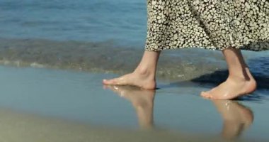 Deniz kıyısındaki kumsalda çıplak ayaklı bir kadının yakından çekimi. Yavaş hareket eden kadın bacakları kıyı şeridinde küçük dalgalar halinde yürür ve su sıçratır. Yüksek kalite 4k görüntü