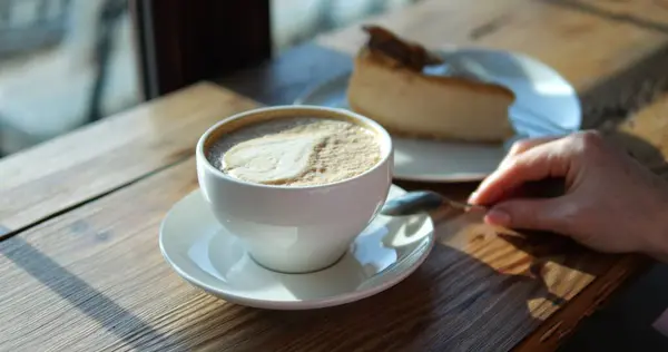 stock image Woman stirs cappuccino with milk froth leaf during breakfast in coffeeshop closeup. Lady with coffee cup and cheesecake at wooden counter. Cappuccino drink