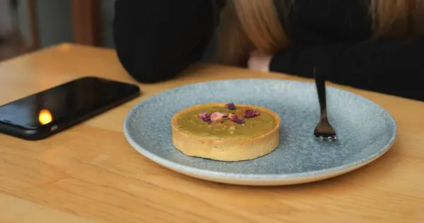 Fıstıklı tart, ahşap masada tabakta ve arkasında da anonim bir kız yatıyor. Yenebilir kurutulmuş çiçeklerle süslenmiş küçük tatlı tart keki. Yanında da akıllı telefon var. 4k görüntü
