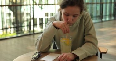 Genç, kafedeki masada otururken samanla keskin narenciye içeceği içiyor. Çocuk, ağır çekim kafenin önünden geçen yayalara karşı soğuk içeceğin tadını çıkarıyor.