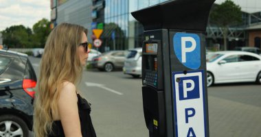 Stylish female driver in sunglasses and black sundress stand at parking meter. Young woman on outdoors parking lot at parked car want to buy parking ticket from parking kiosk. clipart