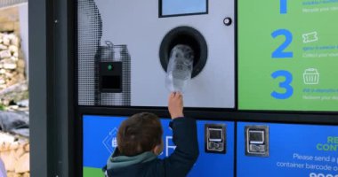 Küçük çocuk plastik şişeyi şehir caddesinde ters satış makinesine sokuyor. Küçük çocuk çevreyi korumak için çöpleri ayırmayı öğreniyor