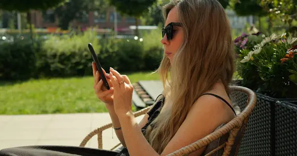 Uzun sarı saçlı, omuz üstü ceketli kadın terasta otururken telefonda mesaj yazıyor. Dijital iletişimin görselleştirilmesi