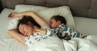 Sevimli küçük oğlu, sakin bir sabahta büyük bir yatakta uyuyan annesine sarılıyor. Rahat kadın ve anaokulu çocuğu yatak odasında yastık ve yorganla uyukluyorlar.