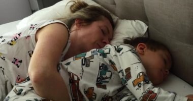 Anne, tembel bir sabahta küçük oğlunu rahat bir yatakta pijama giyerken kucaklıyor. Anaokulu çocuğuyla ilgilenen kadın yatak odasında dinleniyor. Aile eğlencesi.