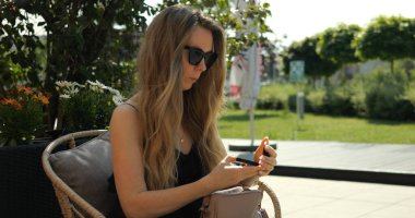 Büyük güneş gözlüklü kadın yaz parkında el çantasından pudra alıyor. Bayan, sokak kafesi terasında dinlenirken çantasında küçük bir ayna buluyor.