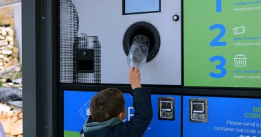 Küçük çocuk plastik şişeyi şehir caddesinde ters satış makinesine sokuyor. Küçük çocuk çevreyi korumak için çöpleri ayırmayı öğreniyor