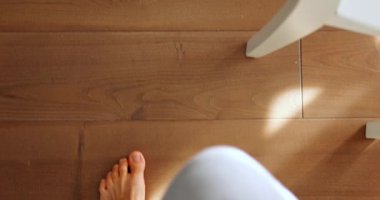 Çıplak ayaklı bir adam parlak güneş ışığıyla ahşap bir odada yürüyor. Açık gri pantolonlu genç adam yavaş çekimde sandalyenin yanından geçen parke üzerinde yürüyor.