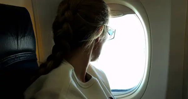 Gözlüklü kadın tatilde, uçak kuaföründe pencereden dışarı bakıyor. Örgülü saçlı kadın yolcu yurt dışına seyahat etmeyi düşünüyor.