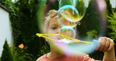 Kıvırcık saçlı çocuk plastik baloncuk asasından bile bir dizi sabun köpüğü salıyor. Komik küçük çocuk kır evinin bahçesinde baloncuk çıkarırken eğleniyor.