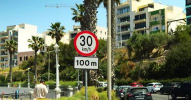 Yol İşareti, işlek caddede, palmiye ağaçlarının arasında yolun bir sonraki bölümü için hızı 30 ile sınırlıyor. Güzergahın yaklaşan kısmı için araçların hareketini azaltan bildirim