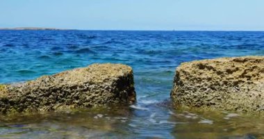 Büyük ıslak kayalara doğru küçük dalgalar kırılır ve sığ sularda köpük oluşur. Büyük yapılar denizde sert doğal oluşumları sembolize eder.