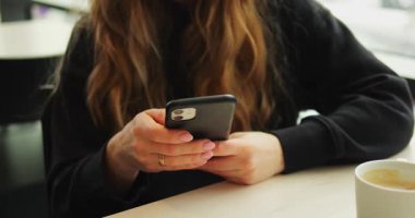 SAVAŞ, POLAND - 10 Mart 2023: dalgalı sarı saçlı kadın telefonu tutuyor ve masada oturan ekrana bakıyor. Sıcak bir fincan kahve yakınlarda durur. Hanımefendinin rahat bir kafede keyfi yerindedir.