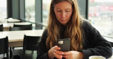 POLAND - 10 Mart 2023: uzun sarı saçlı bir kadın modern kafede oturuyor ve akıllı telefonuyla interneti parşömenliyor. Ekrana odaklanan bir kadının önünde bir fincan kahve.