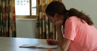 Düşünceli genç bir kadın elinde kalemle not defterine bakıyor. Genç esmer, başını kaldırmış, dirseklerini masaya dayamış.
