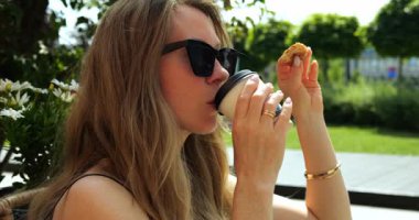 Kadın elinde küçük bir parça hamur işi tutarken tek kullanımlık kahve fincanından bir yudum alıyor. Bayan parkta içkiyle atıştırmayı sever.