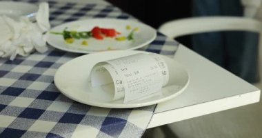 Tabak fiyatları ve toplam miktarı olan bir kağıt parçası masanın üstünde duruyor. Yuvarlanan çek, kantinde yemek prosedürünün sonunu simgeler.
