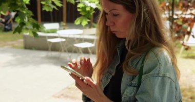 Uzun sarı saçlı bir kadın dışarıda duruyor ve akıllı telefonlara el ele tutuşarak bakıyor. Park ortamında ekran okuma ve yazı yazma üzerine odaklanan bayan