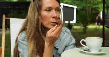 Güneş gözlüklü olgun bir kadın telefon görüşmesi yaptığını belirten akıllı telefonu ağzına kadar tutar. Masada kalan içecekle birlikte fincan, kafede birden fazla görev önerisi sunar