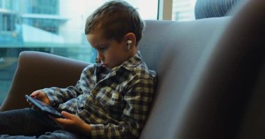 Anaokulu öğrencisi minderli koltuğa oturur ve geniş bir pencere ile el telefonuna odaklanır. Damalı gömlek ve kulaklıklar kafeterya ortamında sakin ve ekran tarafından emilmiş olduğunu gösteriyor