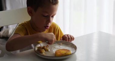 Anaokulu öğrencisi kahvaltıda büyük masada oturur ve peynirli kek yer. Çocuk peynirli krep yer ve tabağa bakarak ekşi kremaya batırır.