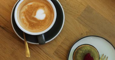 Kalp desenli kapuçino ve ahşap masada küçük tatlı bir pasta. Lüks tatlıyla öğle yemeği ve rahat bir kafede servis edilen porselen bardakta tatlı sıcak içecek.