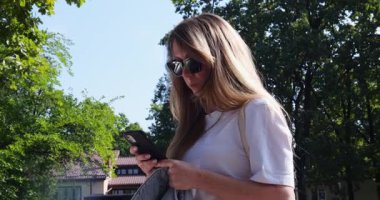 Güneş gözlüklü sarışın kadın yeşil şehir parkında duran akıllı telefonu kontrol ediyor. Beyaz tişörtlü orta yaşlı kadın şehir caddesinde cep telefonu ekranında mesaj atıyor.