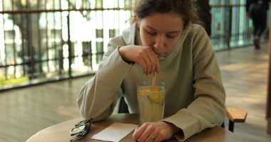 Genç, kafedeki masada otururken samanla keskin narenciye içeceği içiyor. Çocuk, ağır çekim kafenin önünden geçen yayalara karşı soğuk içeceğin tadını çıkarıyor.