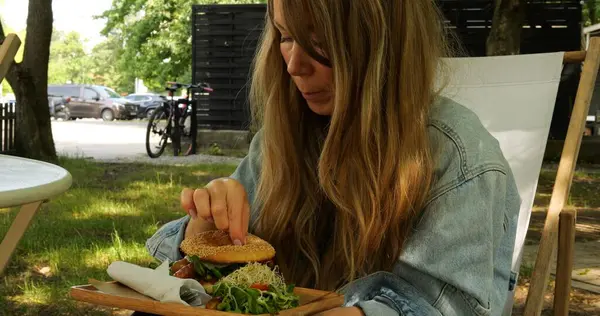 Genç bir kadın, akçaağaç ağaçlarıyla çevrili park kafesinin terasında oturmuş hamburger yiyor. Aç dişiler, sakin doğayla baş başa kalmanın tadını çıkarırlar.