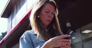 Işıklı sarışın kadın internette akıllı telefonuyla sörf yapıyor süpermarketin önünde duruyor. Yoğunlaşmış kadın sokakta cep telefonuyla mesaj okuyor.