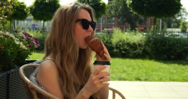 Sarışın kadın kruvasanın tadını çıkarırken tek kullanımlık kahve fincanından kahve içiyor. Bayan, Park Cafe 'de rahatça yemek ve içeceklerle vakit geçiriyor.