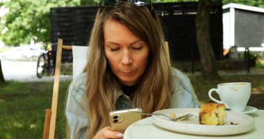 Güneş gözlüklü kadın açık havada masada oturmuş bir fincan kek ve kahveyle telefona bakıyor. Olgun bir kadın telefon etmeye odaklanmış.